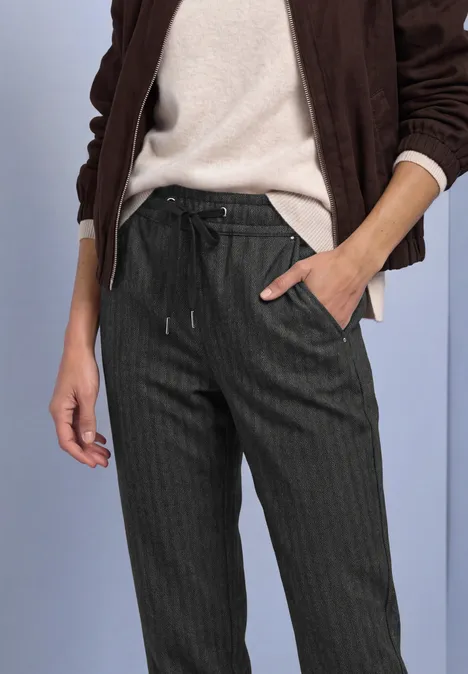 Vrouw in donkere pantalon met fijne strepen en trekkoord, gecombineerd met een lichte trui en jas.