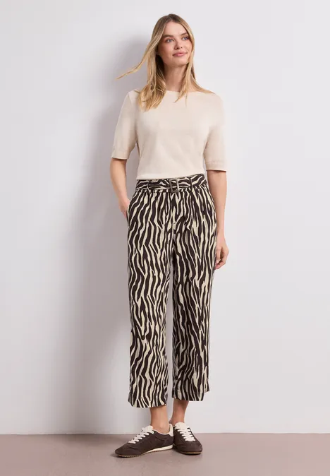 Une personne porte un t-shirt crème à manches courtes et un pantalon ample zébré, avec des chaussures marron.