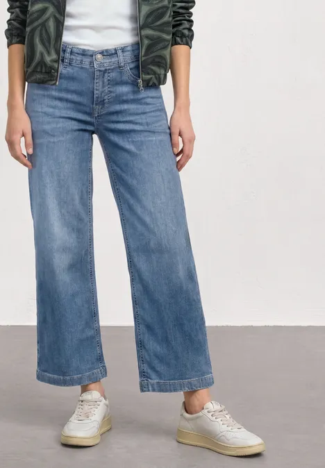 Mid Waist Jeansculotte met elastische tailleband Mid Blue Authentic washed Persoon draagt hooggetailleerde, licht uitlopende, enkellange spijkerbroek in denim met lichte sneakers.