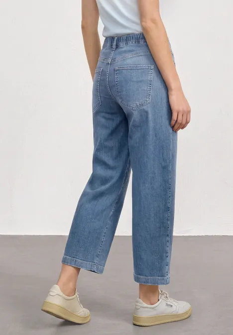 Mid Waist Jeansculotte met elastische tailleband Mid Blue Authentic washed Vrouw van achteren in losvallende, high-waist jeans, lichte sneakers en wit topje tegen een neutrale achtergrond.