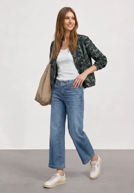 Mid Waist Jeansculotte met elastische tailleband Mid Blue Authentic washed Vrouw in wit T-shirt, donker jasje, blauwe jeans, witte sneakers, draagt tas, tegen neutrale achtergrond.
