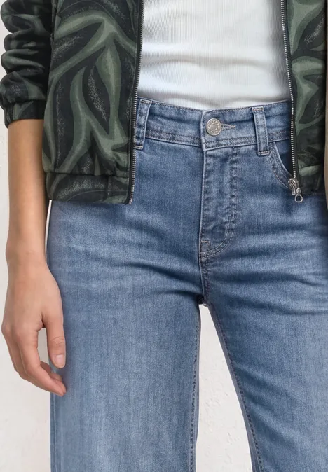 Mid Waist Jeansculotte met elastische tailleband Mid Blue Authentic washed Persoon draagt een high-waist blauwe jeans, een wit T-shirt en een groene jas met camouflageprint.
