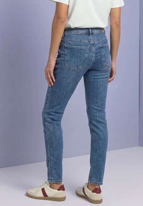 Rückansicht einer Person in schmal geschnittenen blauen Jeans und Sneakern vor hellvioletter Wand.