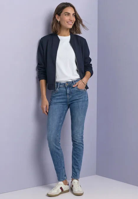 Frau in Slim-Fit-Jeans, weißem T-Shirt, dunkler Jacke und Sneakern vor lila Hintergrund.