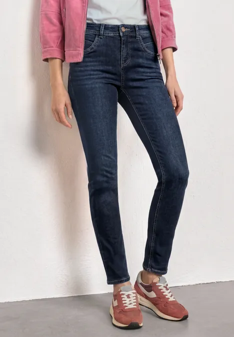 Mid Waist Slim Leg Jeans im Casual Fit Dark Blue Clean washed Frau trägt eng anliegende dunkelblaue Jeans, weiße Bluse, rosafarbene Jacke und rote Sneaker.