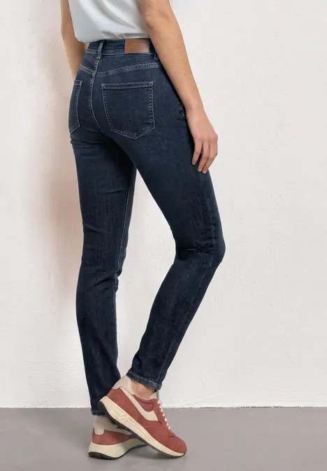 Mid Waist Slim Leg Jeans im Casual Fit Dark Blue Clean washed Eine Person in dunklen Skinny-Jeans und roten Sneakern, von hinten vor einer weißen Wand.