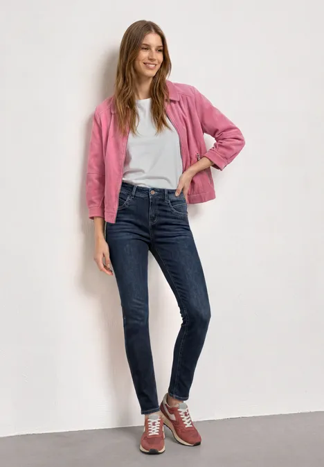 Mid Waist Slim Leg Jeans im Casual Fit Dark Blue Clean washed Frau in pinkem Blazer, weißem Shirt, blauen Jeans und roten Sneakern lehnt locker an der Wand.