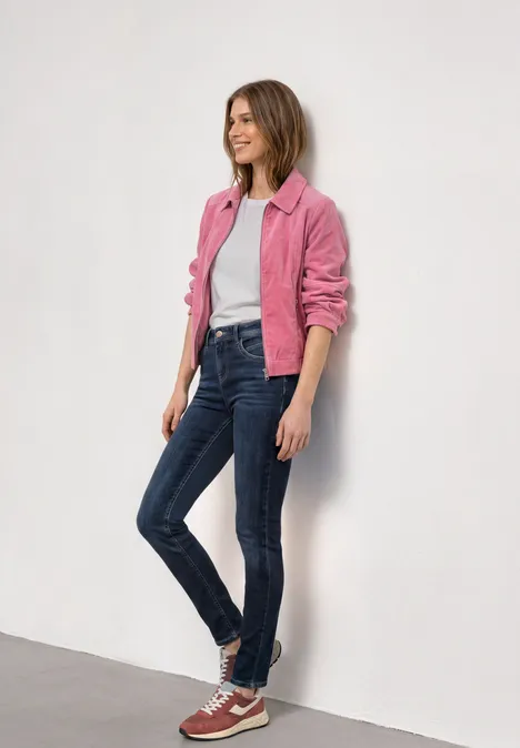 Mid Waist Slim Leg Jeans im Casual Fit Dark Blue Clean washed Eine stehende Person vor weißer Wand in pinker Jacke, weißem Shirt, blauen Jeans und roten Sneakern.