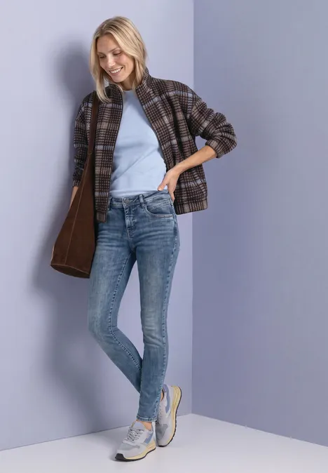 Jeansy Mid Waist z wąskimi nogawkami w stylu Slim Fit Medium Blue Soft Washed Uśmiechnięta kobieta w dżinsach, jasnoniebieskim topie, kraciastej kurtce i trampkach, opiera się o ścianę.