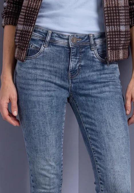 Jeansy Mid Waist z wąskimi nogawkami w stylu Slim Fit Medium Blue Soft Washed Osoba w niebieskich dżinsach, białej koszulce i kraciastej marynarce na neutralnym tle.