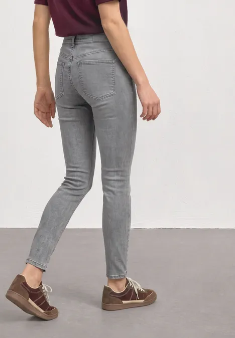 Mid Waist Slim Leg Jeans im Slim Fit Light Grey Random Washed Frau in grauer Skinny-Jeans und braunen Sneakern, von hinten auf neutralem Hintergrund.
