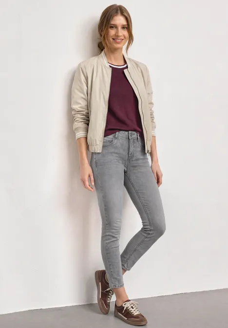 Mid Waist Slim Leg Jeans im Slim Fit Light Grey Random Washed Junge Frau in grauer Jeans, weinrotem T-Shirt und heller Bomberjacke lehnt entspannt an weißer Wand.