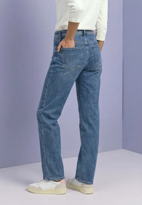 Rückansicht einer Person in geraden blauen Jeans und weißen Sneakern vor lila-grauem Hintergrund.