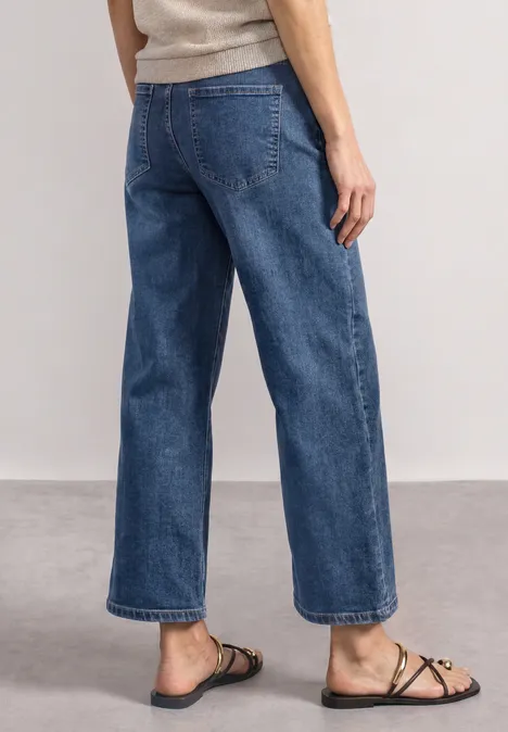 Jeans Culotte mit Gürteldetail Mid Blue Random Washed Schlanke Person trägt weite, cropped Jeanshose und flache Riemchensandalen, stehend auf hellem Boden.