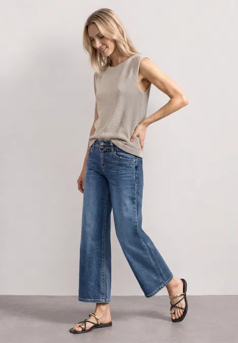 Jeans Culotte mit Gürteldetail Mid Blue Random Washed Frau in grauem ärmellosem Top, weiten blauen Jeans und schwarzen Sandalen vor schlichtem grauem Hintergrund.
