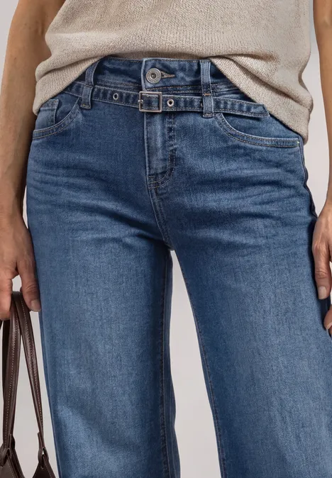 Jeans Culotte mit Gürteldetail Mid Blue Random Washed Eine Person trägt eine beige Bluse und blaue Jeans, hält eine Tasche in der linken Hand.