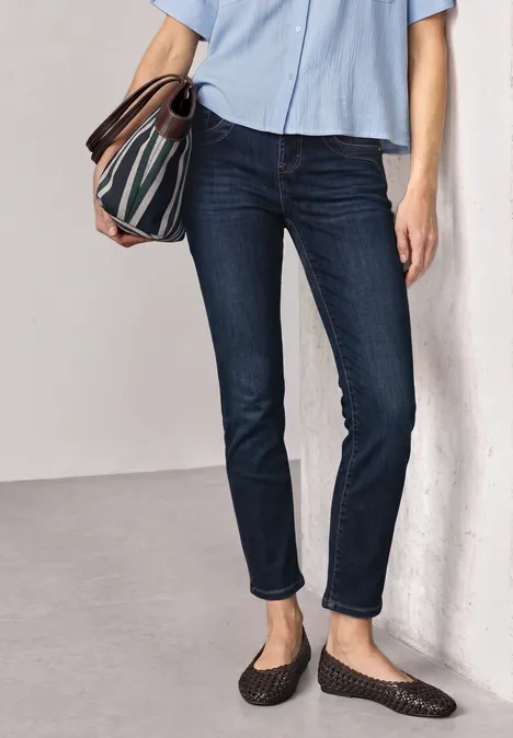 Mid Waist Straight Leg Jeans im Slim Fit Dark Blue Clean washed Person in dunklen Jeans, hellblauer Bluse und gemustertem Umhängetasche, steht entspannt an Wand