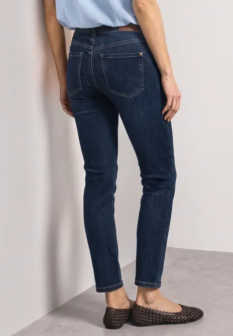 Mid Waist Straight Leg Jeans im Slim Fit Dark Blue Clean washed Rückenansicht einer Frau in dunkelblauen Jeans und flachen, schwarzen Geflecht-Slippern vor einer weißen Wand.