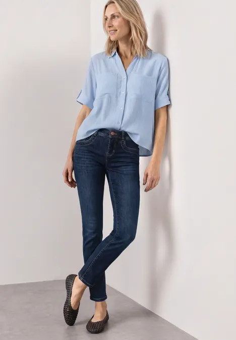 Mid Waist Straight Leg Jeans im Slim Fit Dark Blue Clean washed Frau lehnt an weißer Wand und trägt hellblaue Bluse, dunkle Jeans und schwarze Ballerinas.