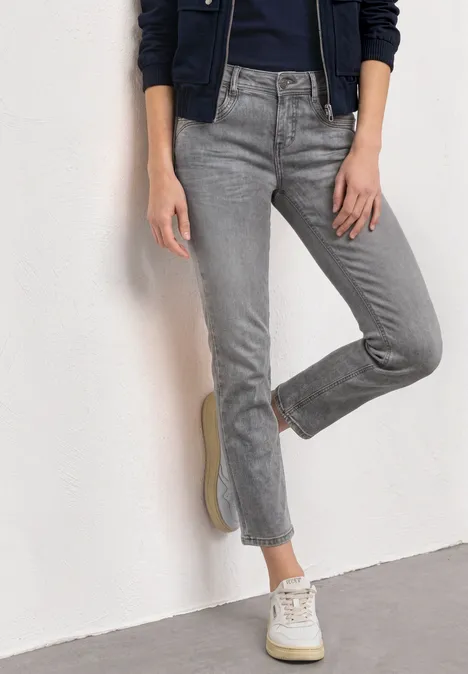 Mid Waist Straight Leg Jeans im Slim Fit Light Grey Random Washed Eine Person steht in grauen, leicht ausgestellten Jeans und weißen Sneakern vor einer weißen Wand.