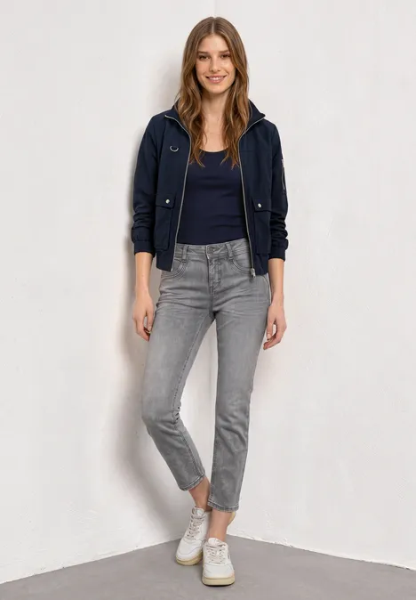 Mid Waist Straight Leg Jeans im Slim Fit Light Grey Random Washed Frau mit dunkelblauer Jacke, schwarzem Shirt, grauen Jeans und weißen Sneakern vor weißem Hintergrund.