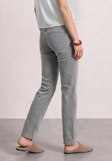 Mid Waist Straight Leg Jeans im Casual Fit Light Grey Random Washed Person von hinten in grauen, geraden Hosen, beigen Hausschuhen und weißem Oberteil.