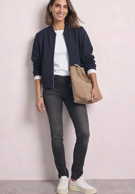 Vrouw in zwarte bomber, wit shirt, grijze jeans, witte sneakers en beige tas voor lichte achtergrond.