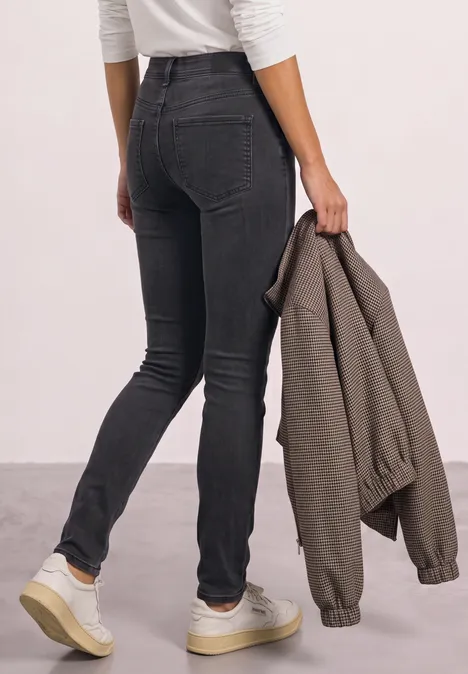 Persoon van achteren in zwarte skinny jeans, witte sneakers aan en een kaki jas in de hand.