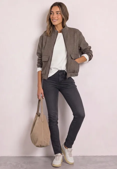 Vrouw draagt bomberjack, wit shirt, zwarte jeans en witte sneakers en houdt een beige tas.