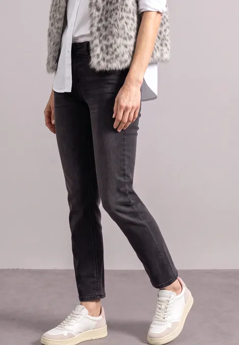 Persoon in zwarte jeans, wit overhemd, grijze nepbontgilet en witte sneakers, neutrale achtergrond.