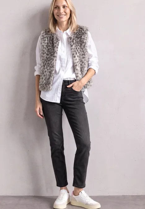Vrouw in witte blouse, zwarte broek, grijze faux fur bodywarmer en witte sneakers voor een lichtgrijze muur.