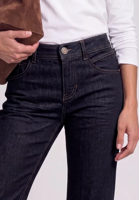 Persoon draagt een witte longsleeve en hoge donkerblauwe jeans, houdt een bruine suède tas vast.