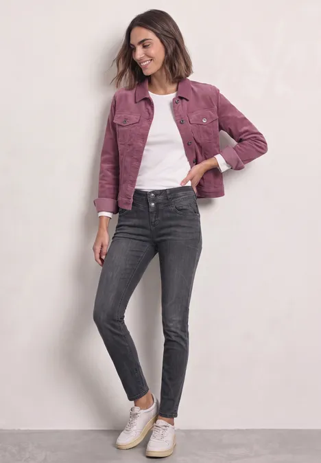 Mid Waist Slim Leg Jeans im Casual Fit Steel Grey Wash Junge Frau in pinker Jacke, weißem T-Shirt, dunkelgrauen Jeans und weißen Sneakern vor hellem Hintergrund.