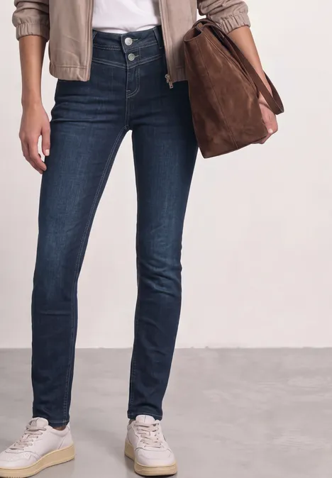 Eine Person trägt dunkelblaue Skinny Jeans, weiße Sneaker, eine beige Jacke und hält eine braune Tasche.