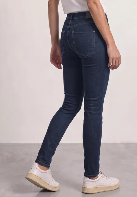 Rückansicht einer Person in dunklen Skinny-Jeans und weißen Sneakern vor neutralem Hintergrund.