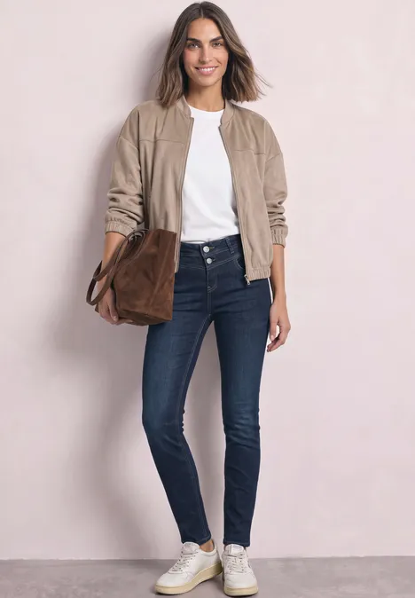Gut gekleidete Frau in Jeans, weißem T-Shirt, beigem Blouson, weißen Sneakern und brauner Tasche vor hellem Hintergrund.