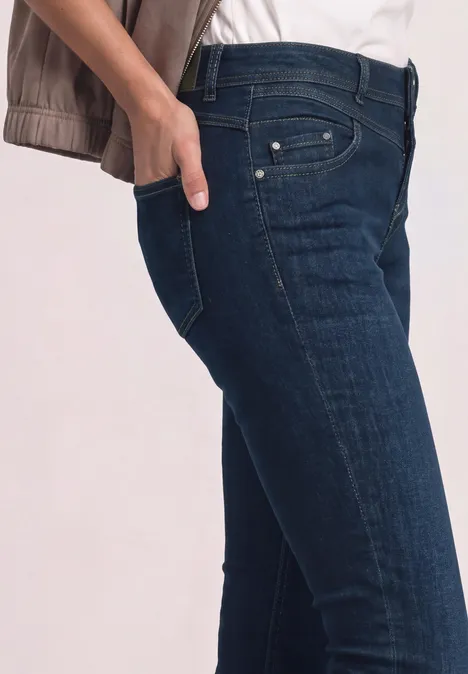 Seitliche Ansicht einer Person in dunklen Jeans, mit Hand in der Hosentasche vor hellem Hintergrund.