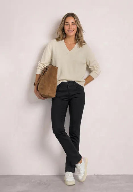 Une femme souriante porte un pull crème, un pantalon noir, des baskets blanches et tient un sac marron.