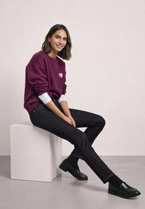 Jean taille moyenne, coupe Slim Leg, style décontracté Deep Rinse Washed Lächelnde Frau sitzt auf weißem Würfel, trägt violettes Sweatshirt, dunkle Jeans und schwarze Loafers vor hellem Hintergrund.