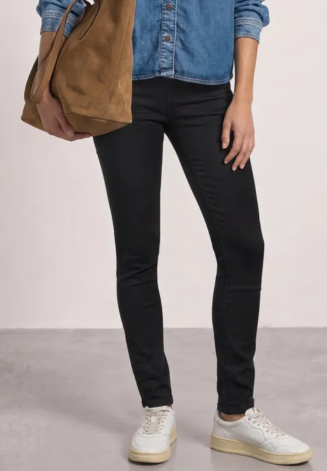 Person in schwarzer Skinny-Jeans, Jeanshemd, weiße Sneaker, trägt olivfarbene Tasche.