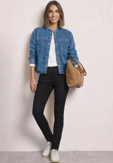 Frau trägt Jeansjacke, weißes Shirt, schwarze Hose, weiße Sneaker und hält eine braune Tasche.