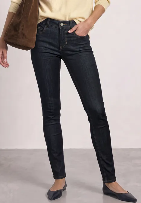 Jean taille moyenne Slim Leg, coupe slim Deep Rinse Washed Dame in dunklen Skinny-Jeans und spitzen Ballerinas, mit beigem Oberteil und Schultertasche.