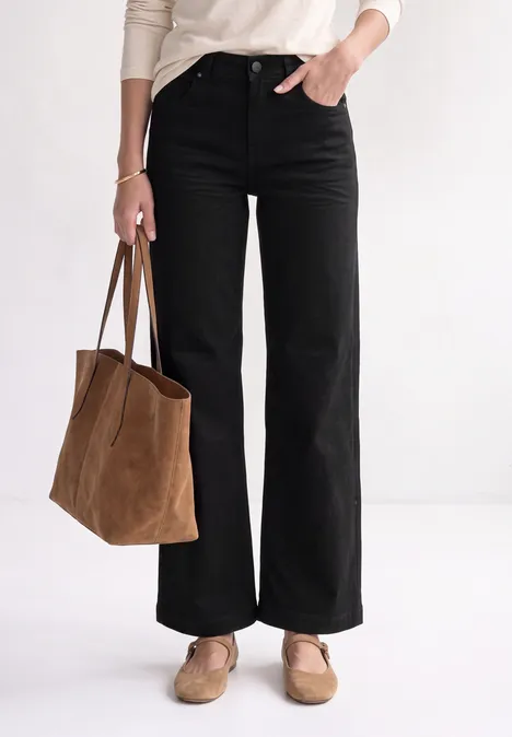Mid Waist Wide Leg Jeans im Loose Fit Black Clean Washed Frau von der Taille abwärts: weite schwarze Hose, weißes Oberteil, beige Loafers und große braune Wildledertasche.