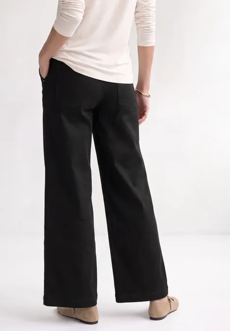 Mid Waist Wide Leg Jeans im Loose Fit Black Clean Washed Person von hinten und seitlich in schwarzer Schlaghose, weißem Oberteil und beigen Schuhen.