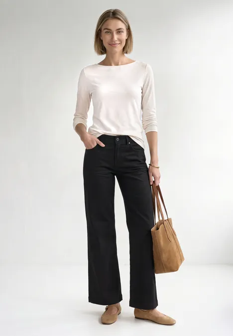 Mid Waist Wide Leg Jeans im Loose Fit Black Clean Washed Frau in weißem Shirt, schwarzen, weiten Hosen, trägt braune Loafers und eine beige Tasche, lächelt.