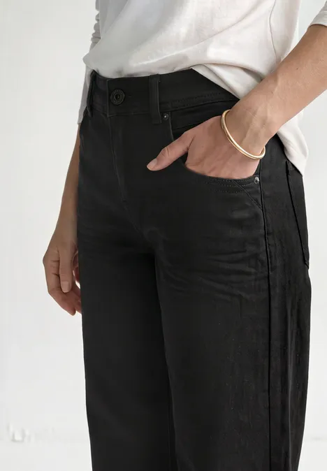 Mid Waist Wide Leg Jeans im Loose Fit Black Clean Washed Schlichtes Outfit mit schwarzen Jeans und weißem T-Shirt, Hand steckt in Hosentasche.