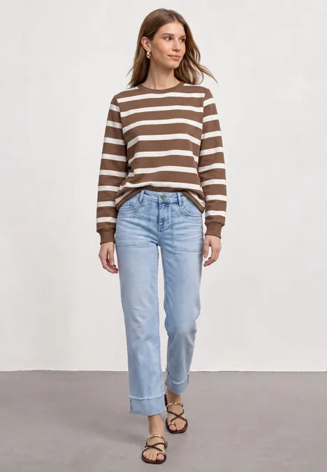Vrouw in bruin-wit gestreepte trui, blauwe jeans en sandalen voor een lichte achtergrond.