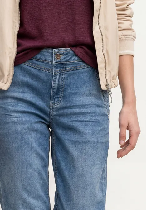 Person trägt blaue Jeans, weinrotes Oberteil und helle Jacke, Hand locker an der Seite.