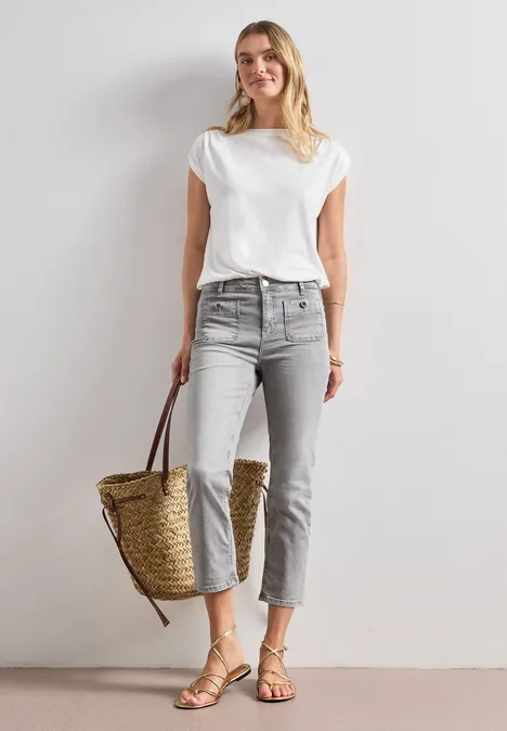 Frau steht in lässigem Outfit aus weißem T-Shirt, grauen Jeans und Sandalen, hält einen Strohkorb.
