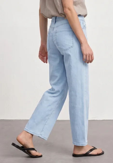 Zijaanzicht van een persoon met lichtblauwe, rechte jeans en zwarte teenslippers.